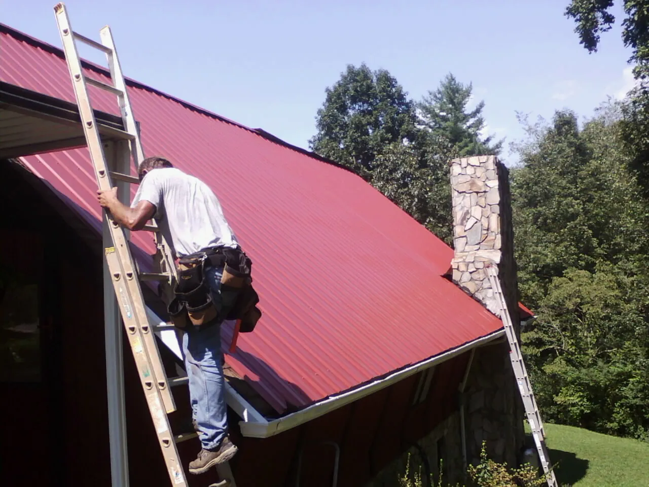 Roofer on a ladder beside a red metal roof and stone chimney — Soffit & Fascia Repair by Roofing Brothers in Duffield